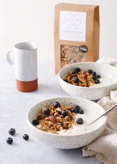 two bowls filled with granola and blueberries next to a bag of granola