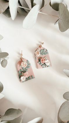 two pairs of pink and green floral earrings on top of white fabric with leaves in the background