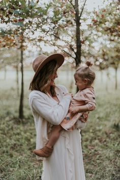 a woman holding a baby in her arms and wearing a brown hat with trees in the background