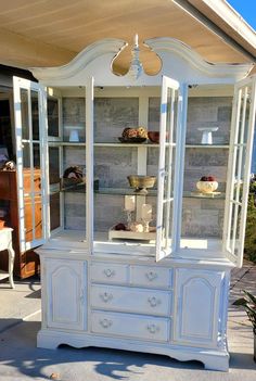 a white china cabinet with glass doors and drawers on the top, in front of a house