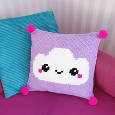 a purple pillow sitting on top of a bed next to a blue pillow with pink pom poms