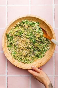 a person holding a wooden bowl filled with green food on top of a pink tile wall