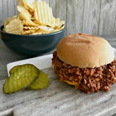 a sloppy joe sandwich next to a bowl of chips and pickle wedges on a marble tray