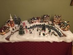 a christmas display with miniature buildings and trees on top of the snow covered tablecloth