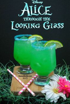 two glasses filled with green liquid sitting on top of a cutting board next to flowers