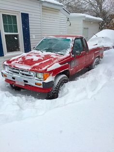 a red pick up truck parked in the snow next to a white house with blue shutters