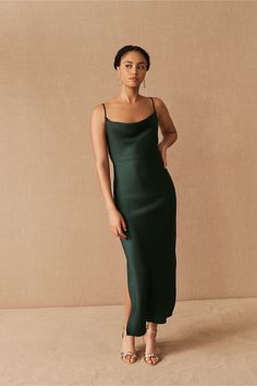 a woman in a dark green dress stands against a beige background and looks off to the side