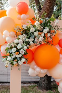 an arrangement of balloons, flowers and greenery in front of a white fence with trees