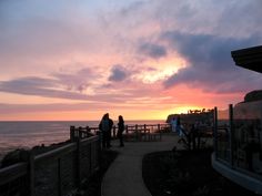 two people are walking on a boardwalk near the ocean at sunset or sunrise with clouds in the sky