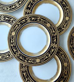 five black and gold plates with designs on them sitting next to each other in front of a white table cloth