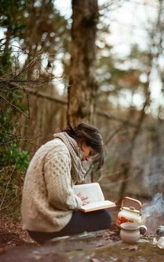 a woman sitting on the ground reading a book next to a campfire in the woods