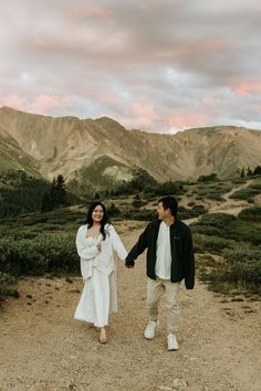 a man and woman holding hands walking down a dirt road in front of some mountains