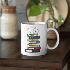 a white coffee mug with a cat sitting on top of some books next to a potted plant