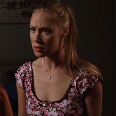 a woman with blonde hair wearing a floral top and looking at the camera while standing in a dark room