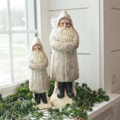 two ceramic santa figurines sitting on top of a window sill with greenery