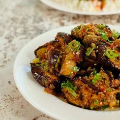 a white plate topped with eggplant covered in seasoning next to a bowl of rice