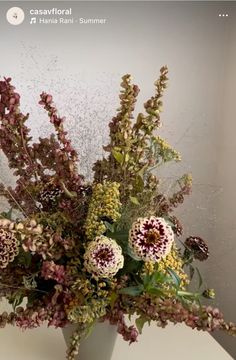 a white vase filled with lots of flowers on top of a table next to a wall