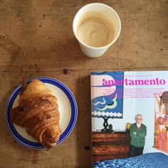 a croissant on a plate next to a cup of coffee and a magazine