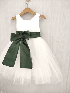 a white and green flower girl dress hanging on a wooden hanger with a green bow