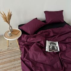 a bed covered in purple sheets and pillows with a magazine on the end table next to it