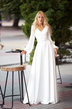 a woman in a long white dress standing next to a table and chair with her hands on her hips