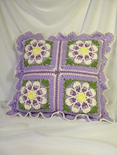 a crocheted purple and green pillow with white flowers on the front, sitting on a white sheet