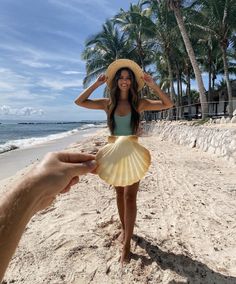 a woman is standing on the beach wearing a hat and holding her hand up to her head