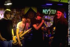 group of young men standing in front of a bar playing guitars and singing into microphones