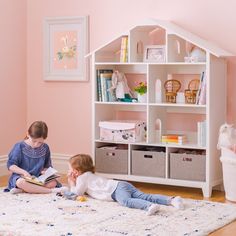 two children are sitting on the floor in front of a doll house and bookshelf