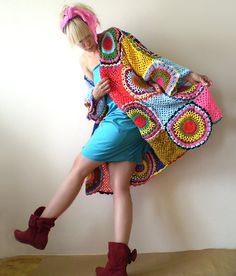 a woman in a colorful crocheted jacket and boots is leaning against a wall