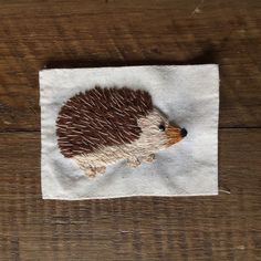 an embroidered hedgehog on a piece of white linen with brown and tan trim, sitting on a wooden surface