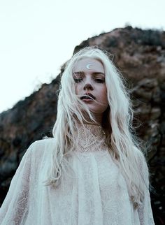 a woman with long white hair standing in front of a rock formation and looking at the camera