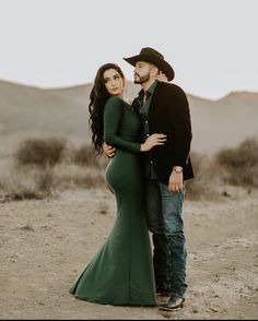 a pregnant woman in a long green dress standing next to a man wearing a cowboy hat