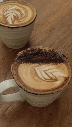 two cups of coffee sitting on top of a wooden table next to each other with designs in them