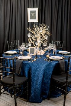 the table is set with black chairs and blue cloths, silver place settings and gold accents