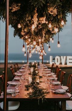 a long wooden table with lots of lights hanging from it's ceiling and surrounded by greenery