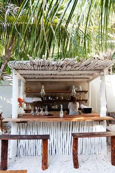 a man sitting at a bar in the sand under a palm leaf covered roof with bottles and glasses on it