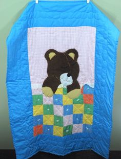 a teddy bear sitting on top of a blue quilted wall hanging from a hook