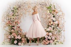 a woman in a pink dress standing next to a floral arch with flowers on it