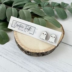 two white ghost earrings sitting on top of a wooden stump next to green leafy branches