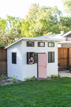 a small white shed with a pink door and window on the side, in front of a fenced yard