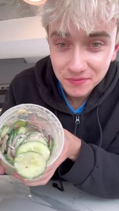 a young man holding up a plastic container filled with cucumbers and other vegetables