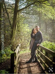 a man and woman standing on a bridge in the woods with their arms around each other