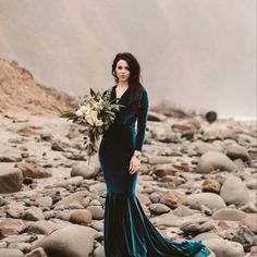 a woman in a long dress standing on rocks holding a bouquet and looking off into the distance