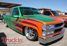 an orange and green truck parked in a parking lot next to other trucks with designs on the hood