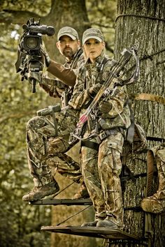 two people are sitting on a tree stand and taking pictures with their camera equipment in the woods