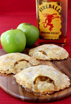 three apple pies on a wooden cutting board next to two green apples and a bottle of fireball cinnamon whiskey