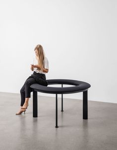 a woman sitting on top of a black circular table next to a white wall and looking at her cell phone