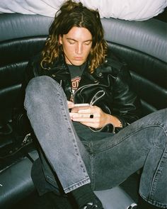 a man with long hair sitting in a chair looking at his cell phone while wearing jeans and a leather jacket