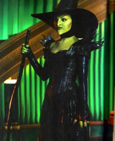 a woman dressed in black holding a cane and wearing a witch costume with green curtains behind her